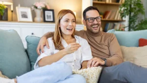 Pariskunta katsoo sohvalla televisiota hymyillen ja syö popcornia